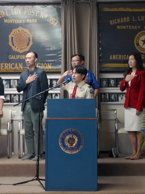 Boy Scout saluting at a podium while three adults stand with hands over their hearts in front of American Legion banners.