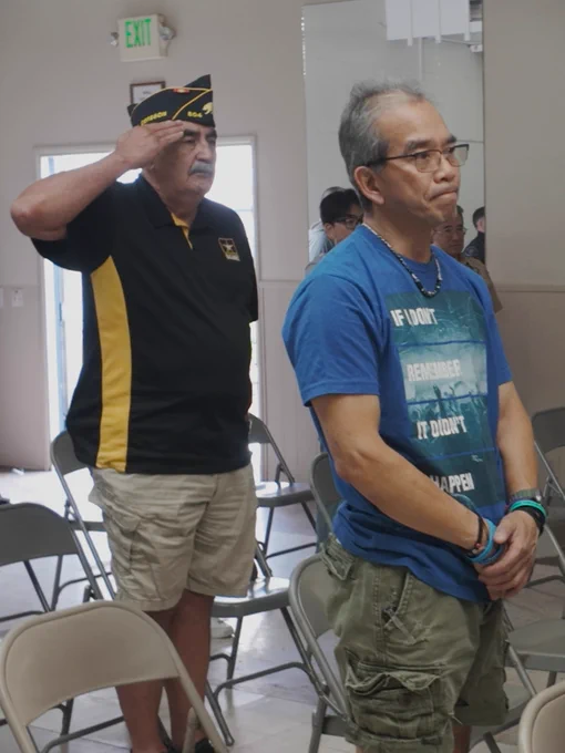 Veteran saluting in a service cap stands behind a solemn man in a blue T-shirt during a ceremony.