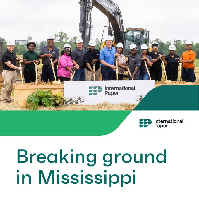 A group of IP employees is standing with shovels at a groundbreaking event in Mississippi, in front of construction equipment. They are posing behind a sign displaying the International Paper logo.