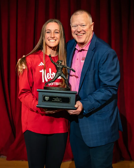 A joyful moment during an award presentation featuring a young woman in a Nebraska softball jersey and a man in a blue blazer holding a trophy together on stage.