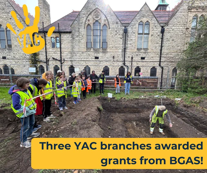 A group of children wearing high-vis vests stand around a pit on an archaeological dig. An archaeologist stands in the pit and is talking to them. It is a cloudy day and historic buildings are in the background. Text in a yellow box says "Three YAC branches awarded grants from BGAS!"