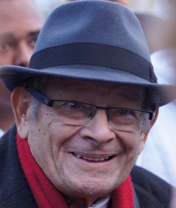 An elderly man wearing a dark fedora hat, black-framed glasses, a black coat, and a red scarf smiles warmly at the camera. The background is blurred, with another person partially visible behind him.