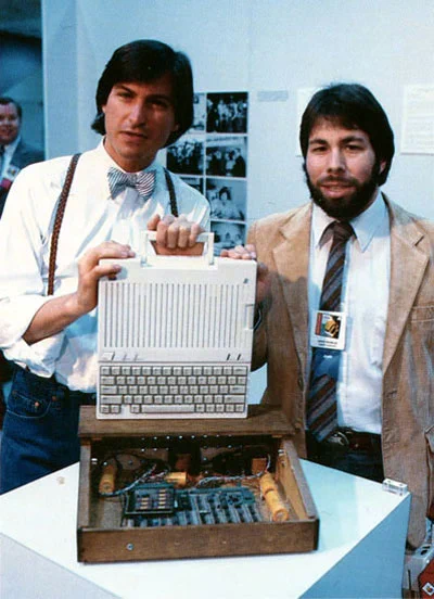 Two men showcase an early computer prototype, with one holding the device and the other standing beside it, highlighting innovation in tech.