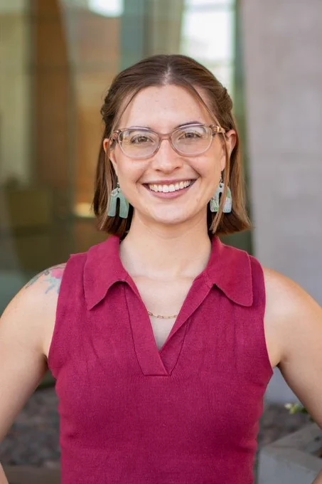 Alexys Murillo wearing a sleeveless dark pink top and turquoise earrings, standing outdoors in front of a blurred building entrance.