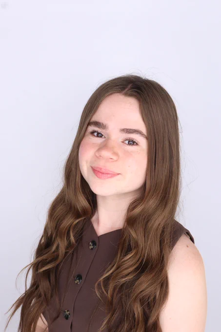 Sophie Byrd smiling slightly with long wavy brown hair, wearing a sleeveless brown button-front top against a light gray background.