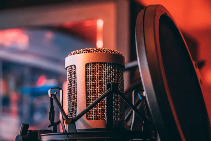 Close-up of a condenser microphone in a shock mount with a pop filter, set in a recording studio with warm red ambient lighting and a softly blurred background.