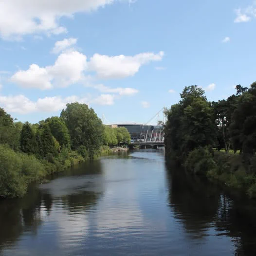 A river flows past tree-lined banks towards a stadium under a cloudy blue sky.