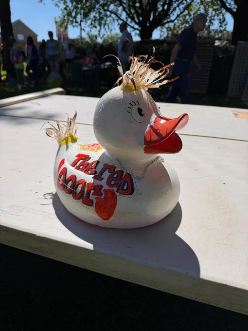 White rubber duck with red beak and feather tuft on head and the words The Red Door on the side, sitting on a white table outdoors in sunlight.