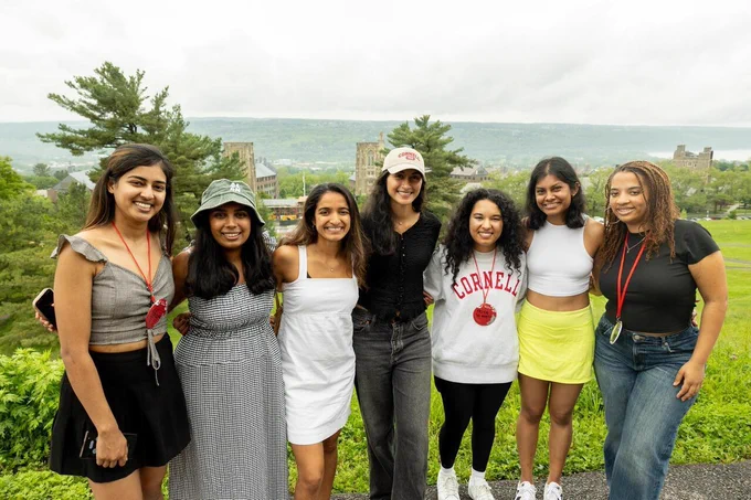 A group of seven people standing outdoors, smiling and posing for a photo. They are casually dressed, with one wearing a shirt that says "Cornell". The background features a scenic view with greenery and distant buildings.