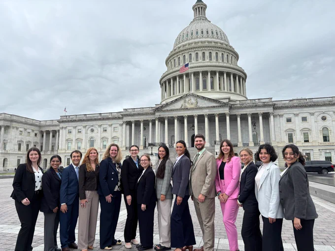 Fourteen individuals (twelve women and two men) representing the 2026 Knauss legislative cohort, posing side-by-side in a single line. The group is dressed in business-professional attire, including black, navy, grey, tan, and pinstripe pantsuits and blazers. One individual stands out on the right side, wearing a vibrant pink pantsuit. They are standing on the stone plaza directly in front of the West Front of the U.S. Capitol Building in Washington, D.C. The iconic white Capitol dome rises prominently behind them, with the American flag flying from the portico below it. The photo was taken during the day under an overcast, heavily clouded gray sky, creating soft, even lighting across the group.
