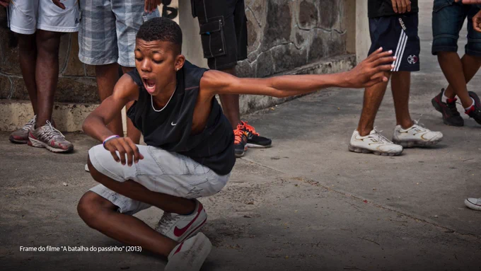 Cena do filme "A batalha do passinho" que destaca um menino negro agachado e dançando. Ele tem o cabelo curto e escuro, a boca aberta, e veste uma regata preta e um shorts branco. Ao fundo, há pernas de pessoas que rondam o menino. 