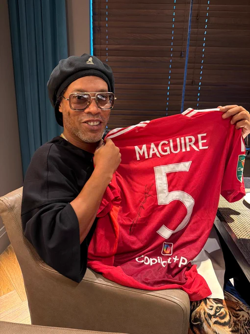 Ronaldinho seated indoors holding a red 2024/25 Manchester United home jersey with the name Maguire and number 5 on the back.