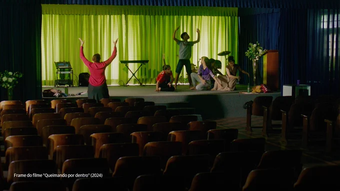 Cena do filme "Queimando por dentro" que destaca um grupo de cinco jovens performando algo em um palco dentro de um auditório. Fora do palco, há uma pessoa de costas para a câmera, de braços esticados para o alto. 