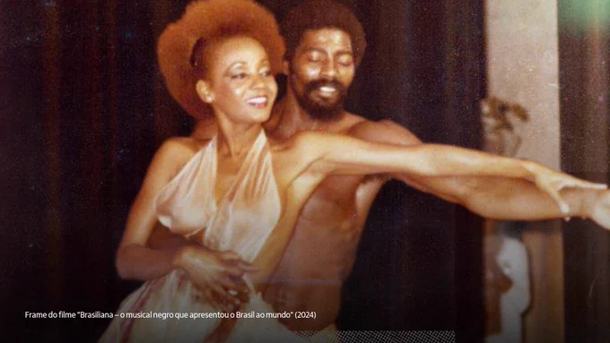 Cena do filme "Brasiliana – o musical negro que apresentou o Brasil ao mundo" que destaca uma mulher e um homem negros sorrindo e dançando. A mulher está na frente e o homem está atrás, ambos esticam o braço esquerdo. A mulher tem o cabelo ruivo e veste uma regata cintilante; e o homem tem cabelo e barba escuros e está com o peitoral nu.