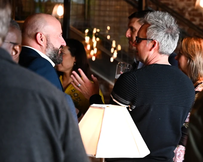 A group of people chat at an indoor social event. Two men are in focus, facing each other and smiling. One holds a drink. Warm lighting and a lamp create a cozy atmosphere. Other guests mingle in the background.