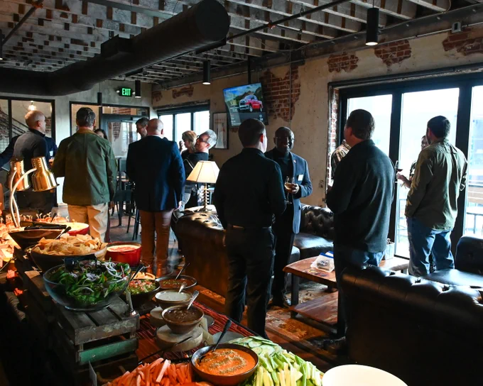 A group of people socialize in a rustic lounge with exposed brick walls, large windows, and a TV. A table in the foreground is filled with appetizers, dips, vegetables, and chips.