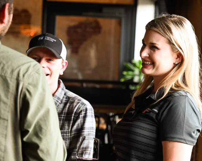 A woman smiling and talking with two men at an indoor social gathering. One man wears a plaid shirt and cap, while the other’s back is to the camera. Warm lighting and a window are visible in the background.