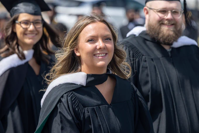 Three smiling graduates in their regalia on a sunny day. 