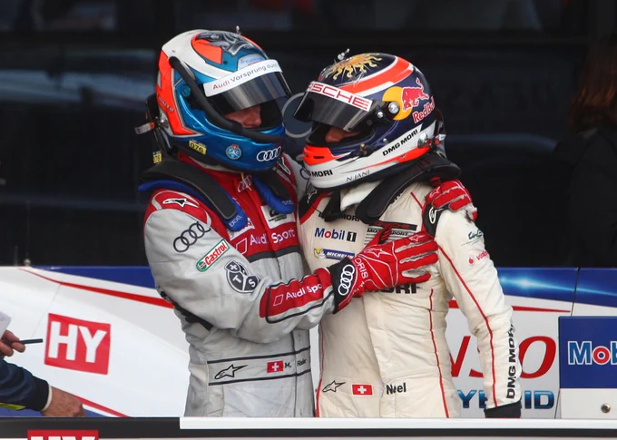 In parc ferme, Marcel Fässler and Neel Jani stand together in a half embrace after steeping out of their respective cars. Fässler, left, wears a red and white race suit and blue and red crash helmet; Jani, right, wears a white race suit and Red Bull sponsored blue and red crash helmet. 2015 6 Hours of Silverstone.