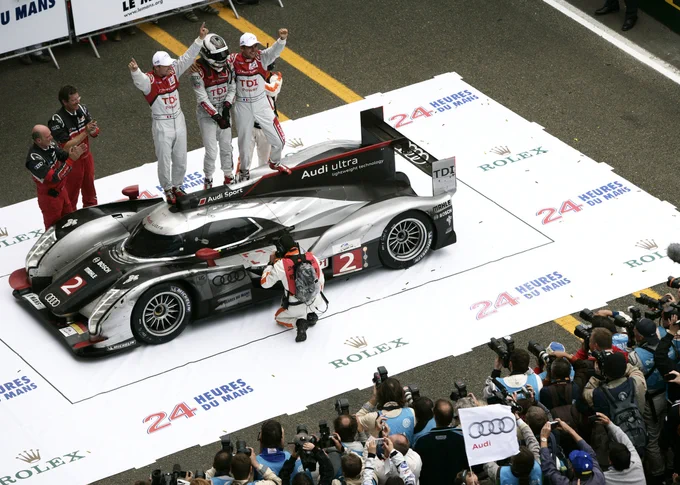 A high angle view shot from the pit lane building of the three Audi drivers - left to right, Marcel Fässler, André Lotterer and Benoît Tréluyer - standing on the roof of their shared Audi R18 TDI in the winner's position in parc ferme. In the bottom of frame, a large number of photographers shoot the scene. 2011 Le Mans 24 Hours.