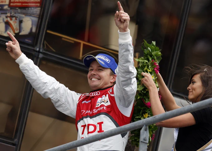 A grinning Marcel Fässler arrives on the walkway to the podium, both arms in the air as he looks out at the crowd below. He wears his red and white Audi race suit and a blue Michelin cap, as a woman wearing a sash readies the winner's laurels for him behind. 2011 Le Mans 24 Hours.