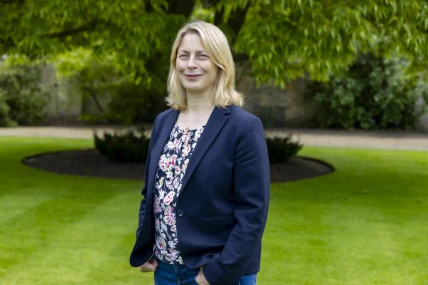 A woman is standing and smiling in a park with green grass and a tree and bushes blurred in the background.