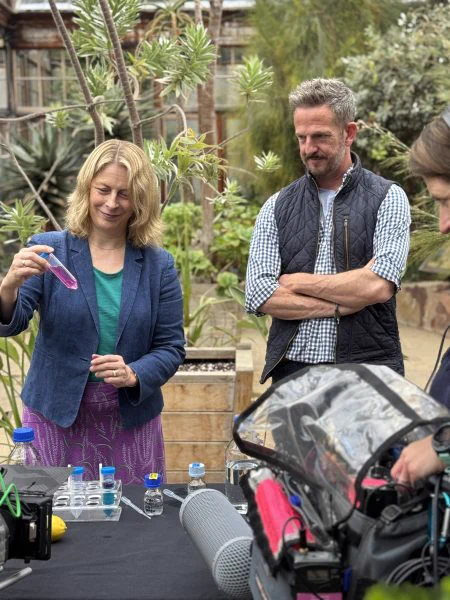 Two people stand behind a table with vials of liquid, pipettes and recording equipment. The woman examines a purple liquid while the man looks on. They are surrounded by Mediterranean plants.