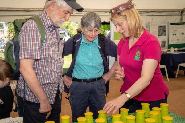 Three people are engaged in a discussion at a table with stacked yellow cups. The person on the right is gesturing towards the cups and wears a T-shirt with "Cambridge University Botanic Garden" on it