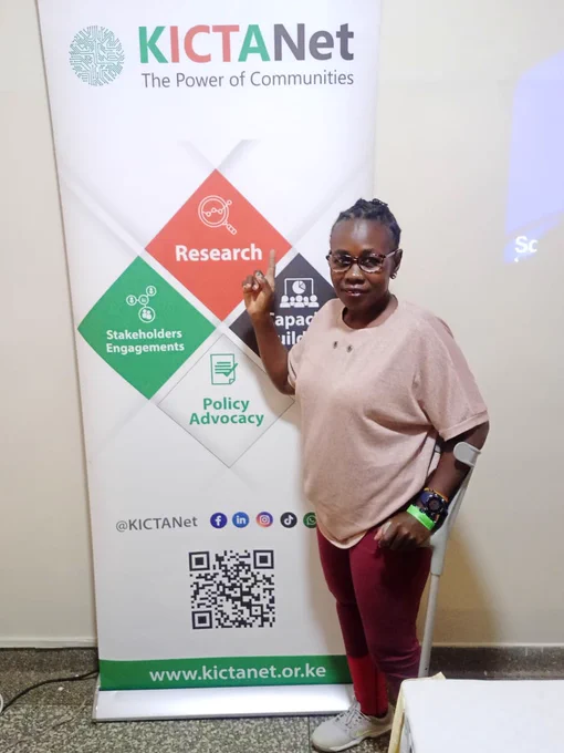 The photo shows a woman standing beside a KICTANet banner. She is smiling and pointing toward the banner, which highlights KICTANet’s key areas of work: Research, Stakeholder Engagements, Capacity Building, and Policy Advocacy. She is dressed casually in a light pink top, maroon leggings, and white sneakers, wearing glasses and a smartwatch. She is also using a crutch.