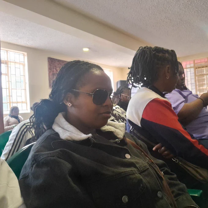 A picture of a woman wearing dark sunglasses and a black denim jacket with a white collar sits calmly, creating a focused and reflective atmosphere. Around her, other attendees are engaged in conversations and activities, with some using their phones and interacting in small groups. 
