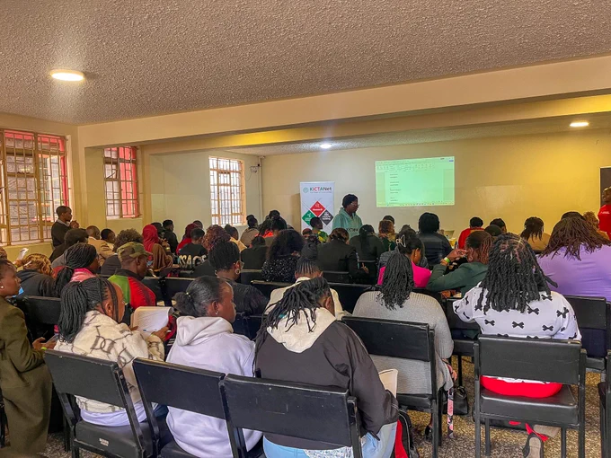 A large gathering during a dialogue session organized in partnership with KICTANet. The room is filled with participants seated attentively and facing the front, where a presentation is being projected onto a screen. A KICTANet banner is visible near the presentation area. Participants are actively engaged, with some taking notes, reading materials, or listening closely to the facilitators.
