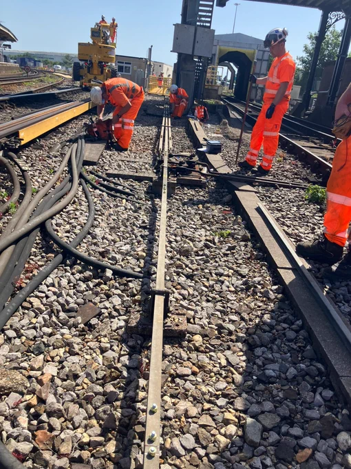 Track engineers in orange PPE are working on track, two using mechanical saws to cut track, and another standing holding a long metal pole