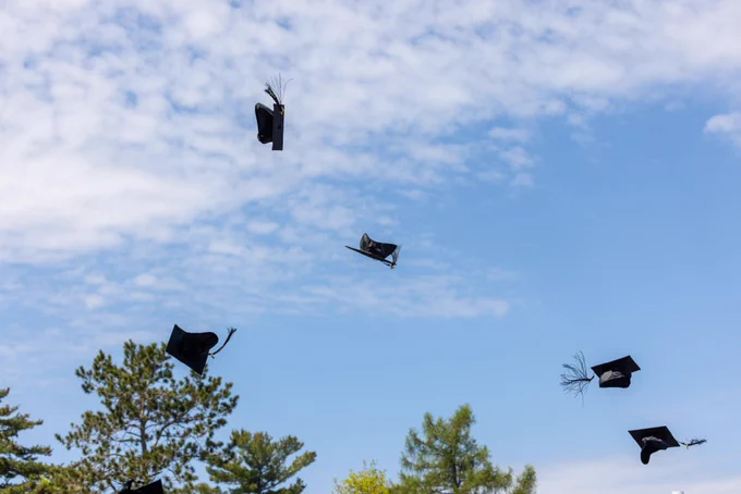 Graduation caps flying in the air against a blue sky with scattered clouds, surrounded by treetops.