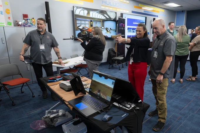 Group of adults in a tech demo room testing virtual reality firearms with laptops and monitors on tables around them.
