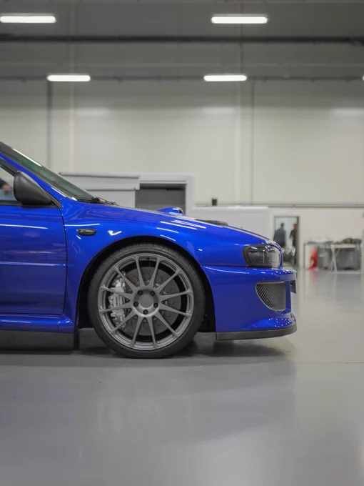 Front quarter view of a blue Prodrive P25 inside the Banbury workshop, showing the classic Subaru Impreza shape, wide arches, multi spoke wheels and large brake setup under bright factory lights