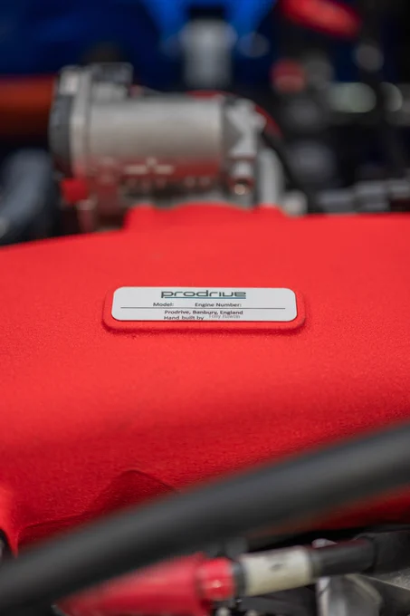 Close up of a Prodrive engine bay with red intake plenum and metal plaque showing hand built details, highlighting craftsmanship and precision engineering inside the P25