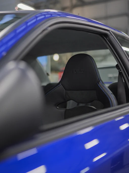 Interior detail, shot through the open window, of the Prodrive P25 showing a black leather bucket seat with P25 embossing, framed by the blue bodywork and workshop setting in the background