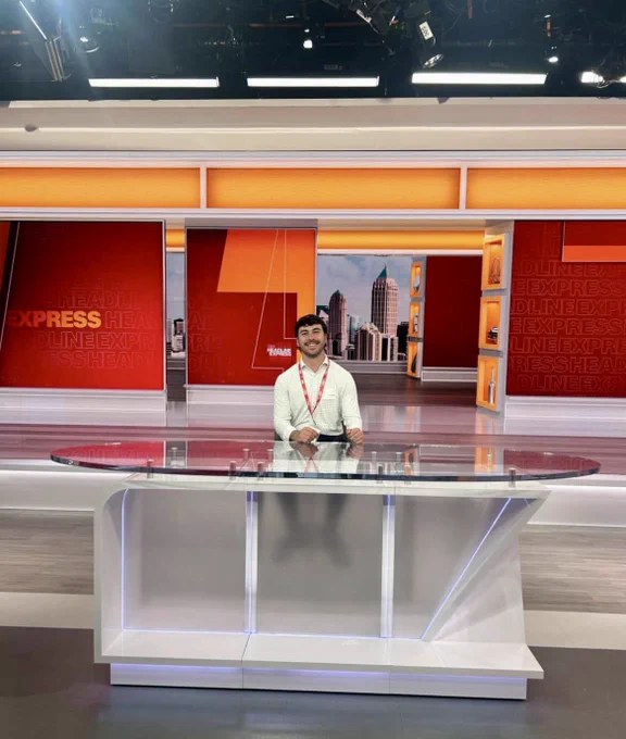 A person standing behind a modern glass news desk in a brightly lit studio with a red and orange background.