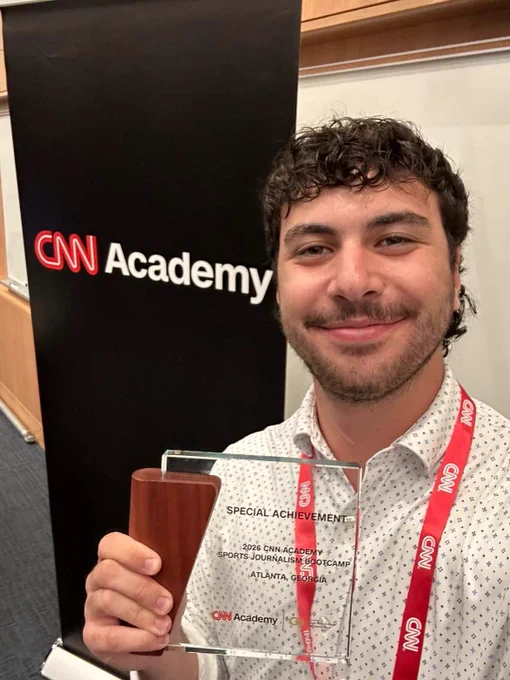 The image shows a person holding a clear rectangular award with a wooden base, standing in front of a large black banner with the "CNN Academy" logo in white and red. The person is smiling and wearing a white shirt with a subtle dot pattern and a red lanyard with "CNN" branding around their neck. The background appears to be an indoor setting, possibly a conference room, with a white board partially visible.