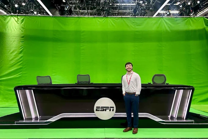 The image shows a person standing beside a sleek, modern anchor desk set against a vivid green screen backdrop. The desk is glossy black with smooth, curved edges and features a prominent circular logo of "ESPN" at the center. Three high-backed office chairs with mesh backs are placed behind the desk. The ceiling is equipped with numerous lights and scaffolding, indicative of a professional studio setup. The green screen is taut and uniformly covers the background, suggesting the setting is intended for broadcasting.