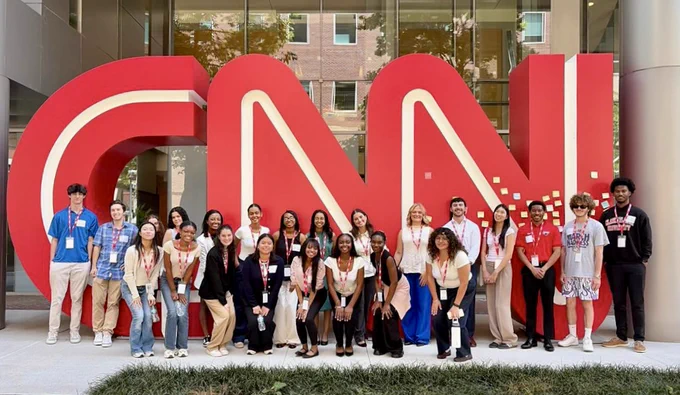 A group of twenty diverse young adults is posing in front of large, three-dimensional red letters spelling "CNN." The sculpture features white lines defining the letters. The group is standing on a stone pavement, surrounded by lush green grass at the foreground. Behind them is the facade of a modern building with glass windows reflecting greenery, suggesting an outdoor setting. The individuals are wearing casual clothing, with a formal stringed identification badge around their necks.