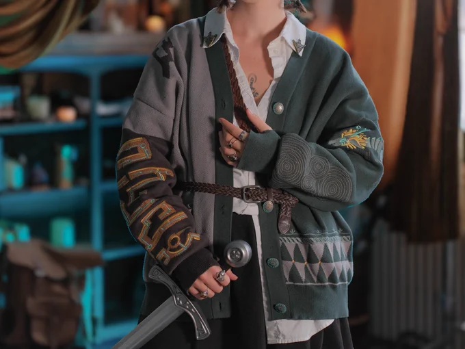 Person standing in a room with blue light behind them wearing an oversized cardigan in shades of teal and grey holding a sword