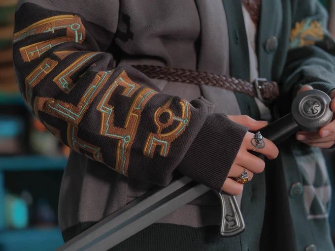 Person standing in a room with blue light behind them wearing an oversized cardigan in shades of teal and grey holding a sword . Close up image of their arm sleeve embroidery
