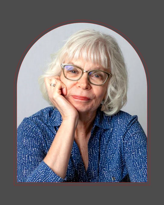 An older white woman with a blue textured top on. She has white hair and glasses and is smiling and resting her face on her right hand.