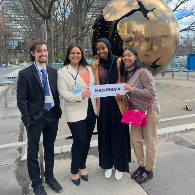 A group of four people smiling, standing outside in front of a large, golden, spherical sculpture. One person holds a sign reading "Micronesia." Trees and a building appear in the background.