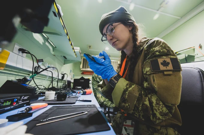 Canadian soldier wearing blue gloves inspects a small electronic device inside a lab.