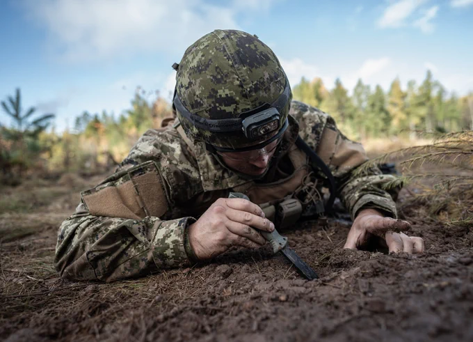 Soldier lying prone on the ground, digging into soil with a tool.