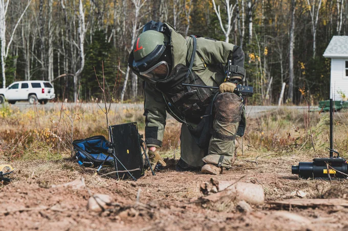 Person in bomb disposal suit kneeling on dirt ground, operating equipment in an outdoor area.