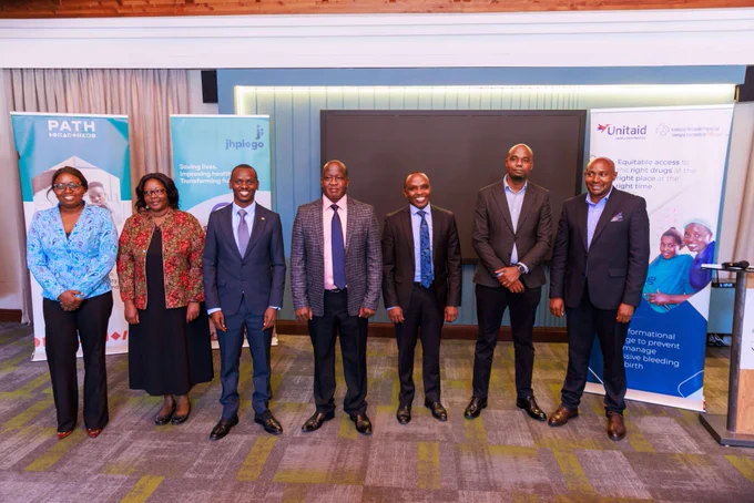 A group of seven individuals stands in a line indoors, dressed in formal attire. Behind them are banners displaying logos and names of organizations such as PATH, Jhpiego, and Unitaid. The setting appears to be a professional event or conference.