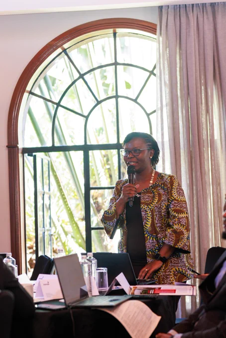 A person stands speaking into a microphone in a room with a large, arched window. The person is wearing a colorful, patterned jacket. There is a table in front with laptops, paperwork, and a water bottle.